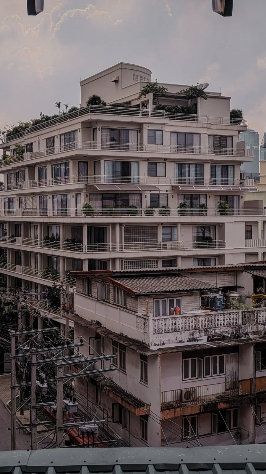 A terraced Bangkok apartment block at dusk, balconies full of potted plants above older shophouse roofs and tangled power lines