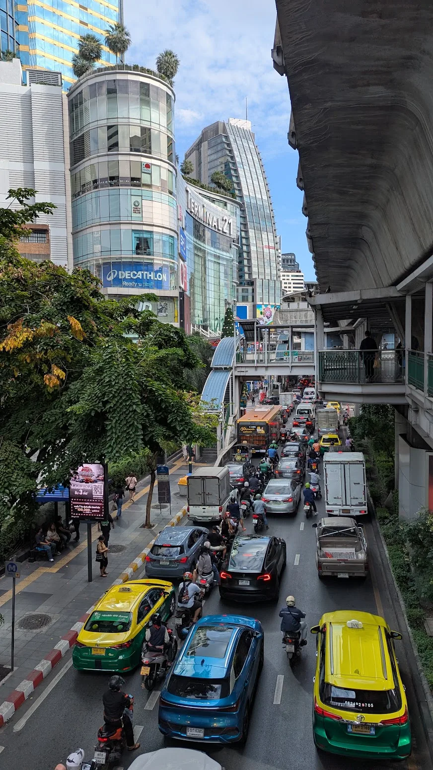 Traffic and motorbikes below the BTS skywalk at Asok, with the Terminal 21 mall rising behind the trees