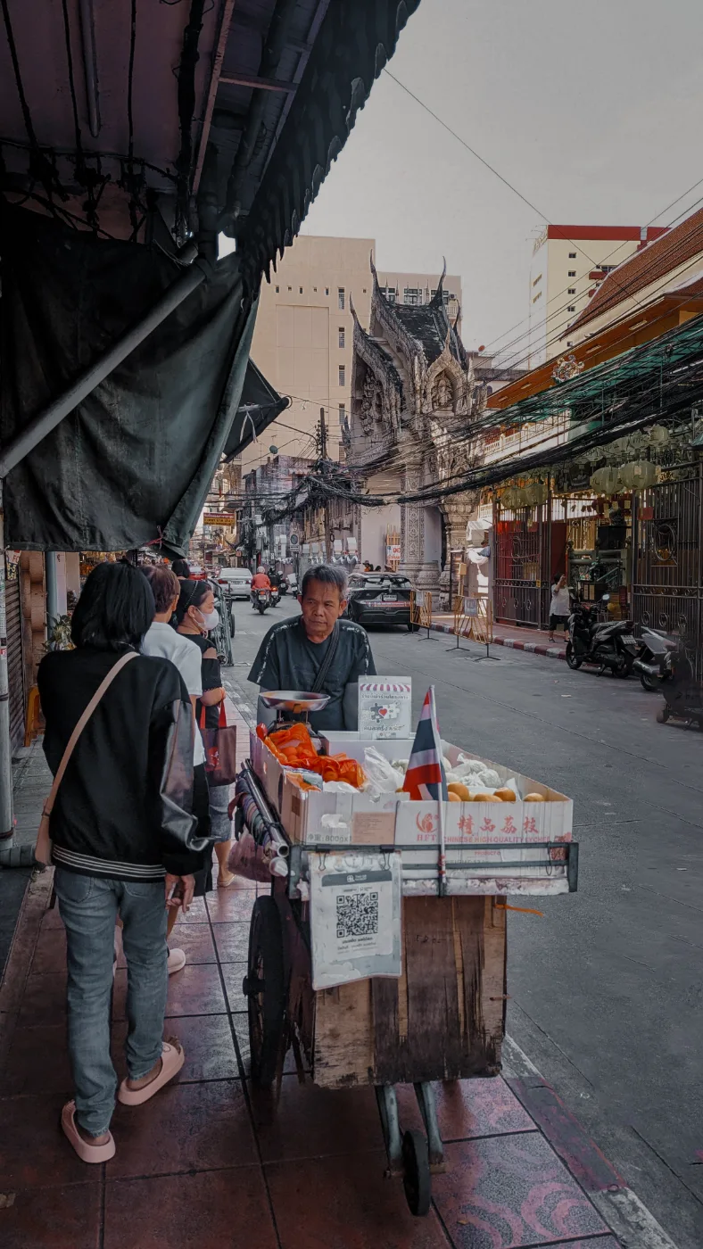 A fruit-cart vendor with a Thai flag on his cart, an ornate temple gable rising behind him on a Chinatown lane