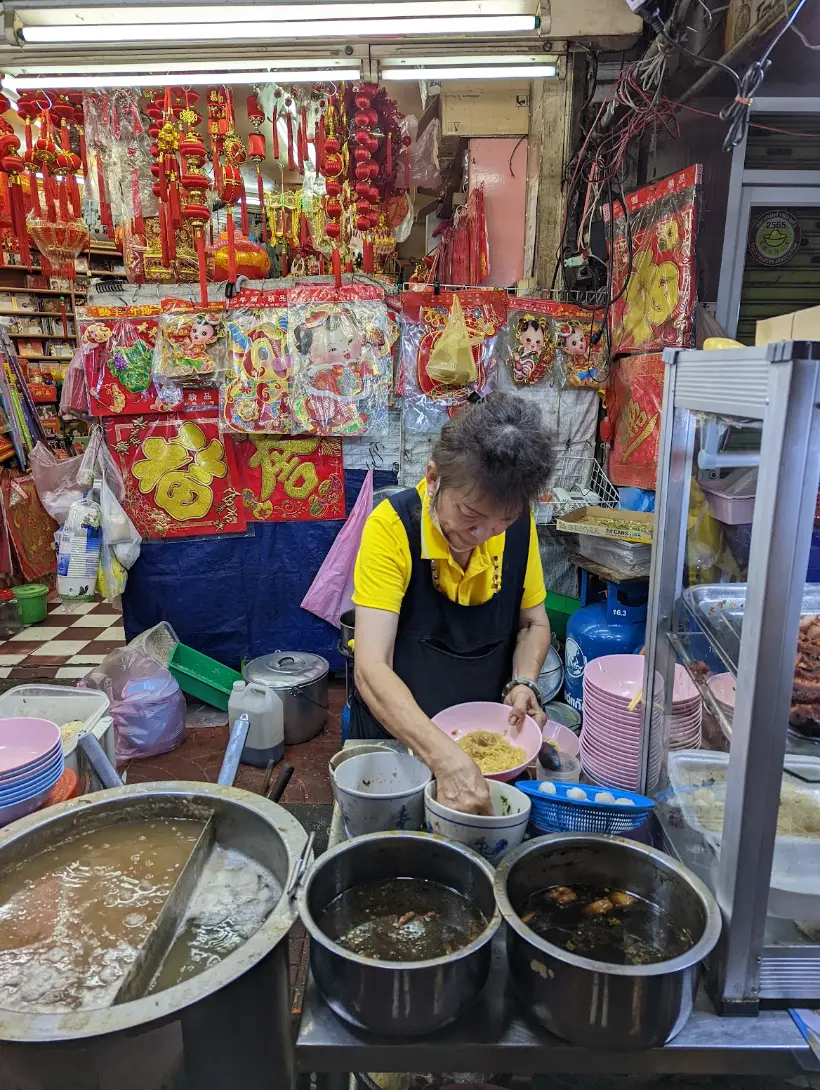 A cook lifting egg noodles into a bowl at a Chinatown street stall, red Lunar New Year decorations hanging above the counter