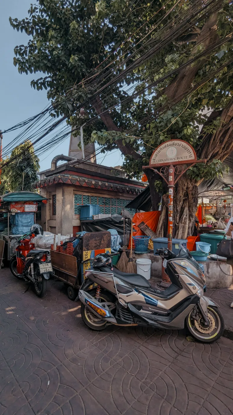 A silver scooter parked under a banyan tree beside a Chinatown Route marker sign, with market crates behind