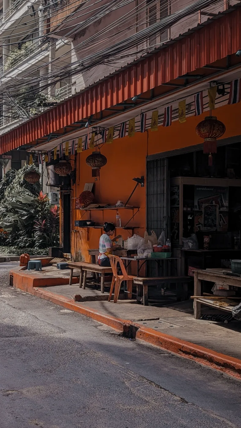 An orange-walled open-front shop under a red corrugated awning on an Asok side street, hung with Thai flags and Chinese lanterns, a woman prepping food at a table out front