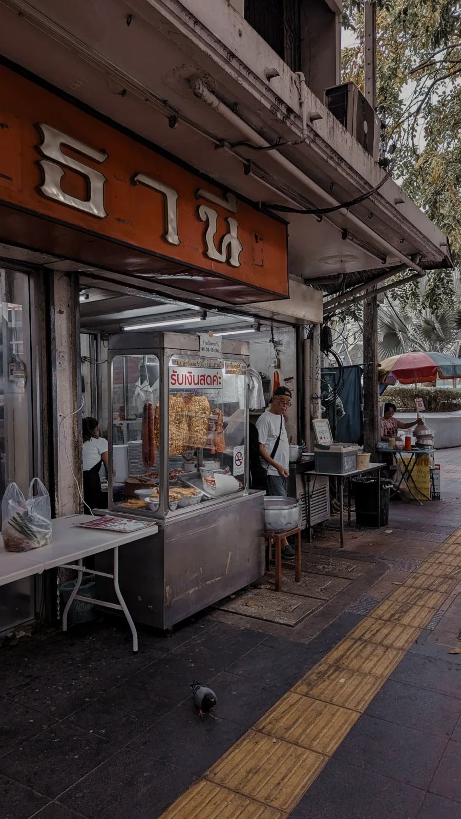 A roast pork shopfront on a quiet Bangkok sidewalk, glass case hung with moo daeng and crispy pork
