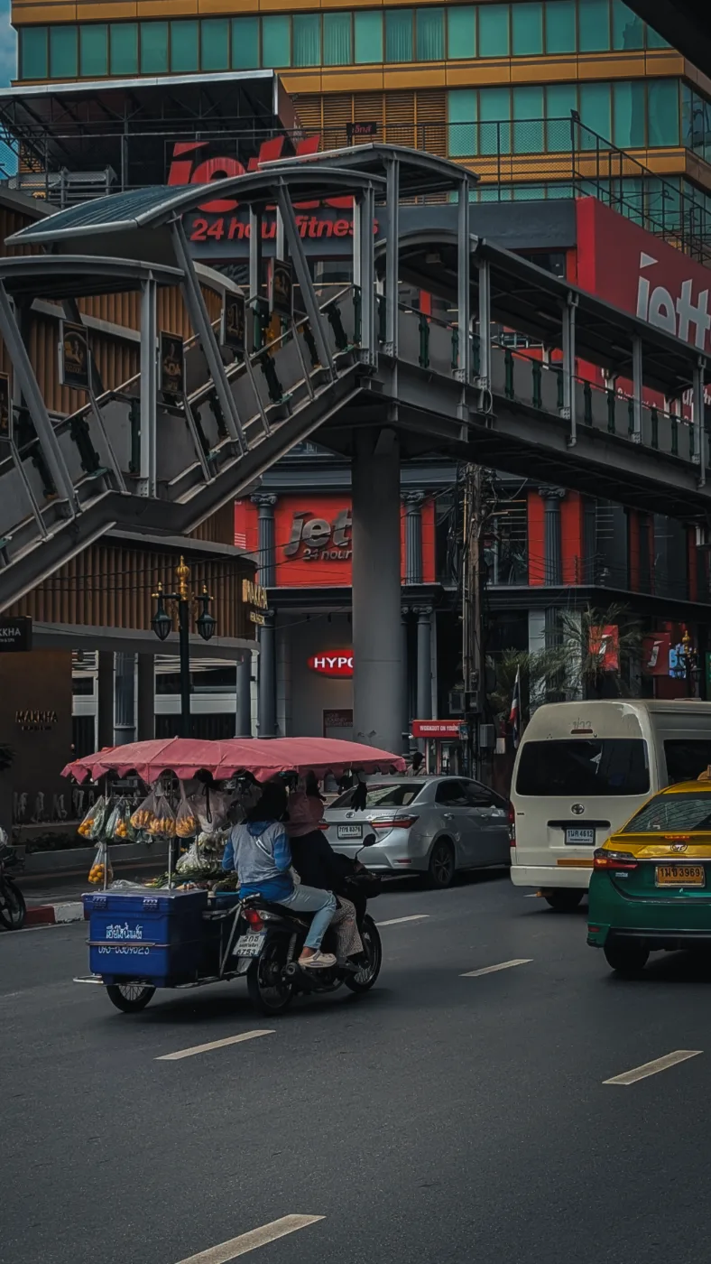 A fruit vendor’s motorbike cart crossing a wide Bangkok road beneath a pedestrian skywalk and lit shopfront signs