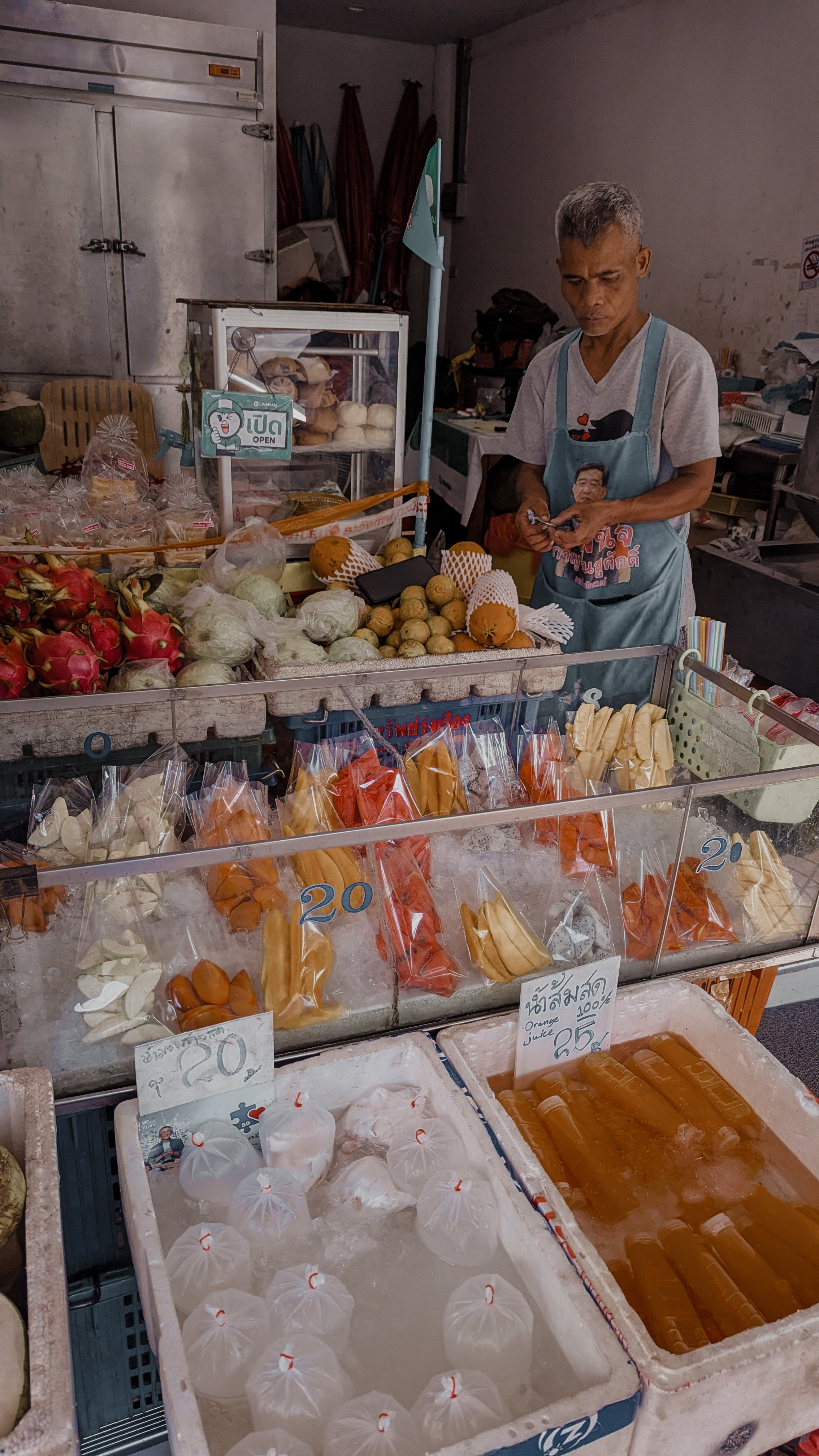 A vendor behind a glass cart of bagged cut fruit and fresh orange juice on ice at a Bangkok market
