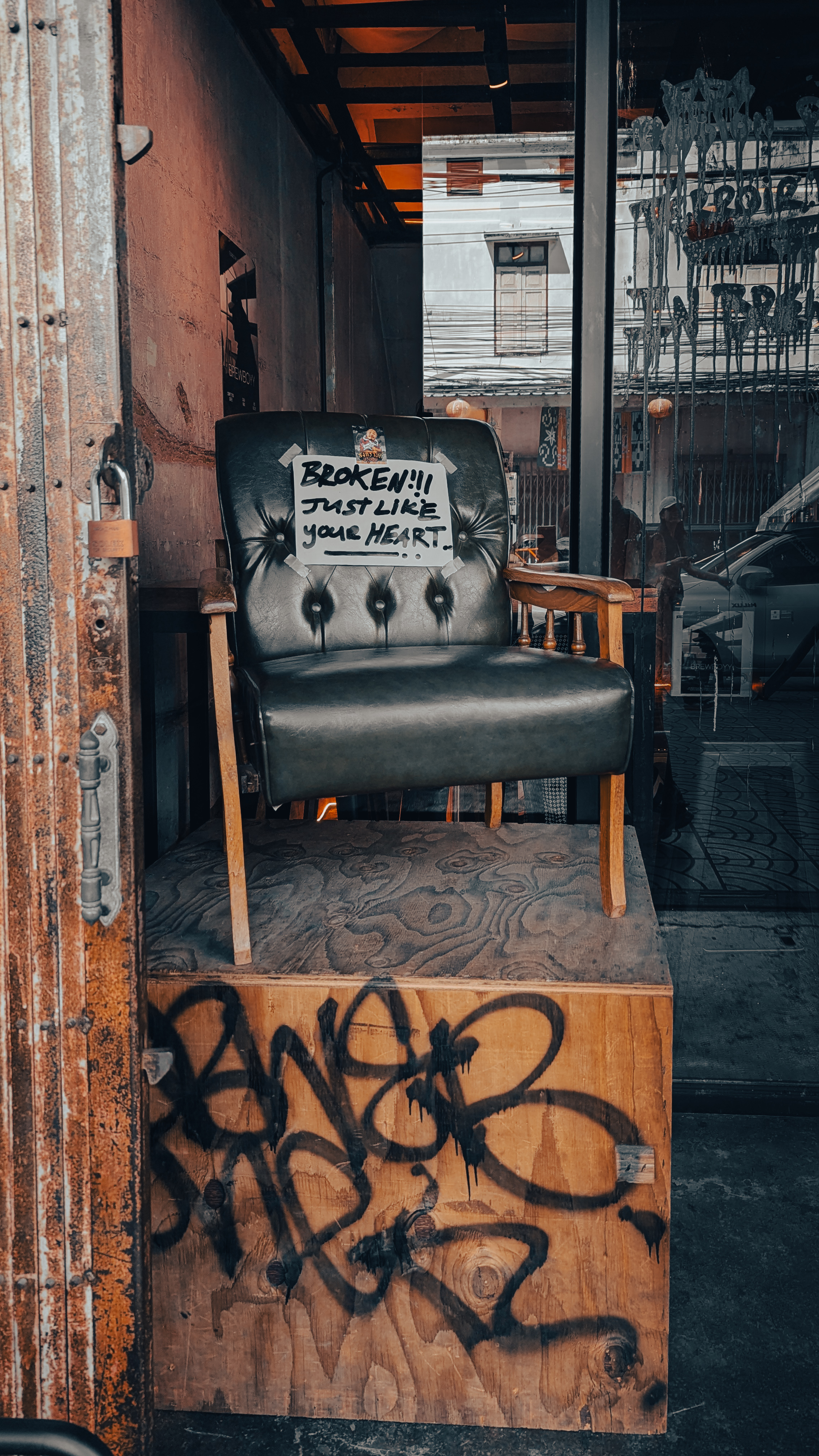 A worn leather armchair on a graffitied plinth outside a Song Wat bar, with a hand-lettered sign reading Broken, just like your heart
