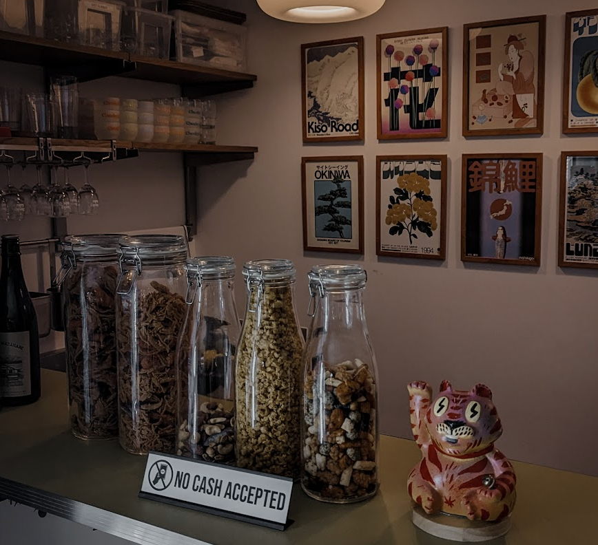 Cheeky Sake bar shelf: glass jars of snacks, a red maneki-neko cat figurine, a ‘No cash accepted’ sign, and a wall of Japanese travel posters