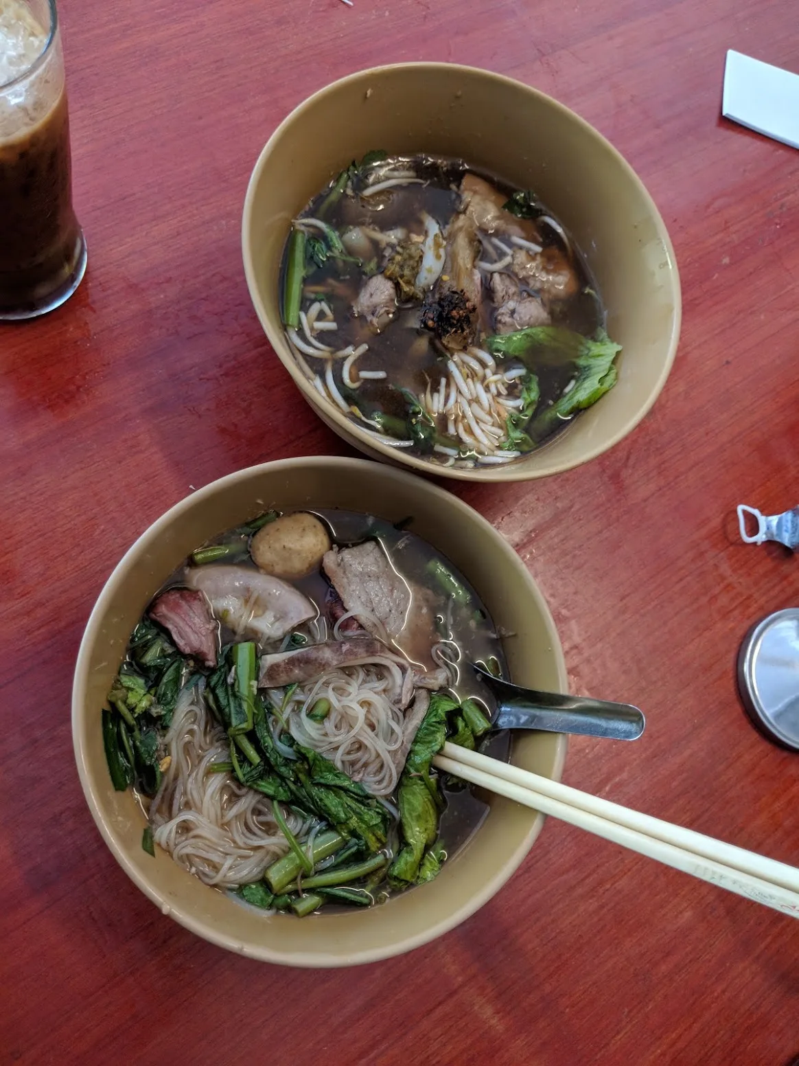 Two bowls of boat noodles with pork and greens beside an iced coffee