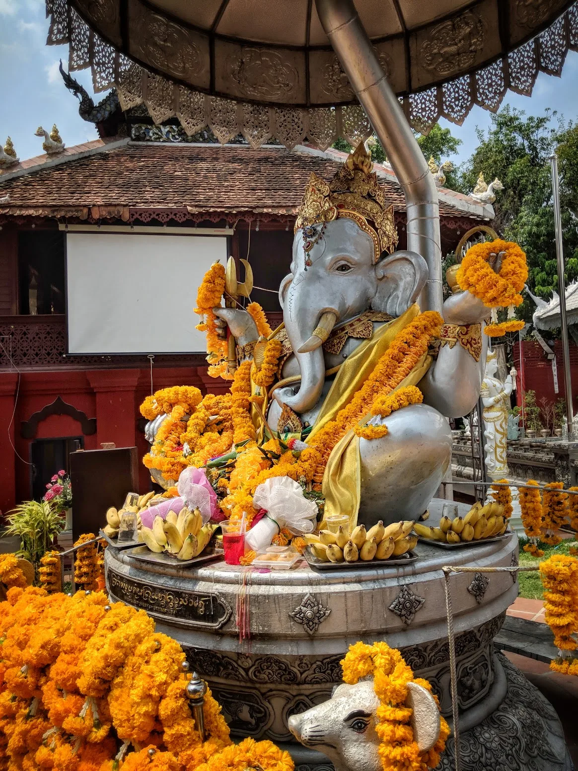 A silver Ganesha shrine under a tiered parasol, draped in marigold garlands with banana offerings at its base