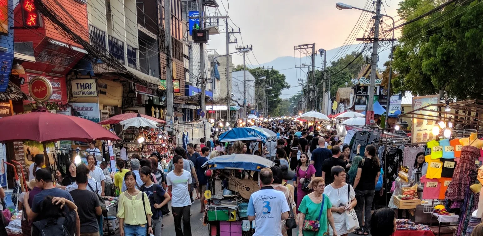 A crowded weekend walking street at dusk: stalls under umbrellas, shoppers filling the road, hills behind