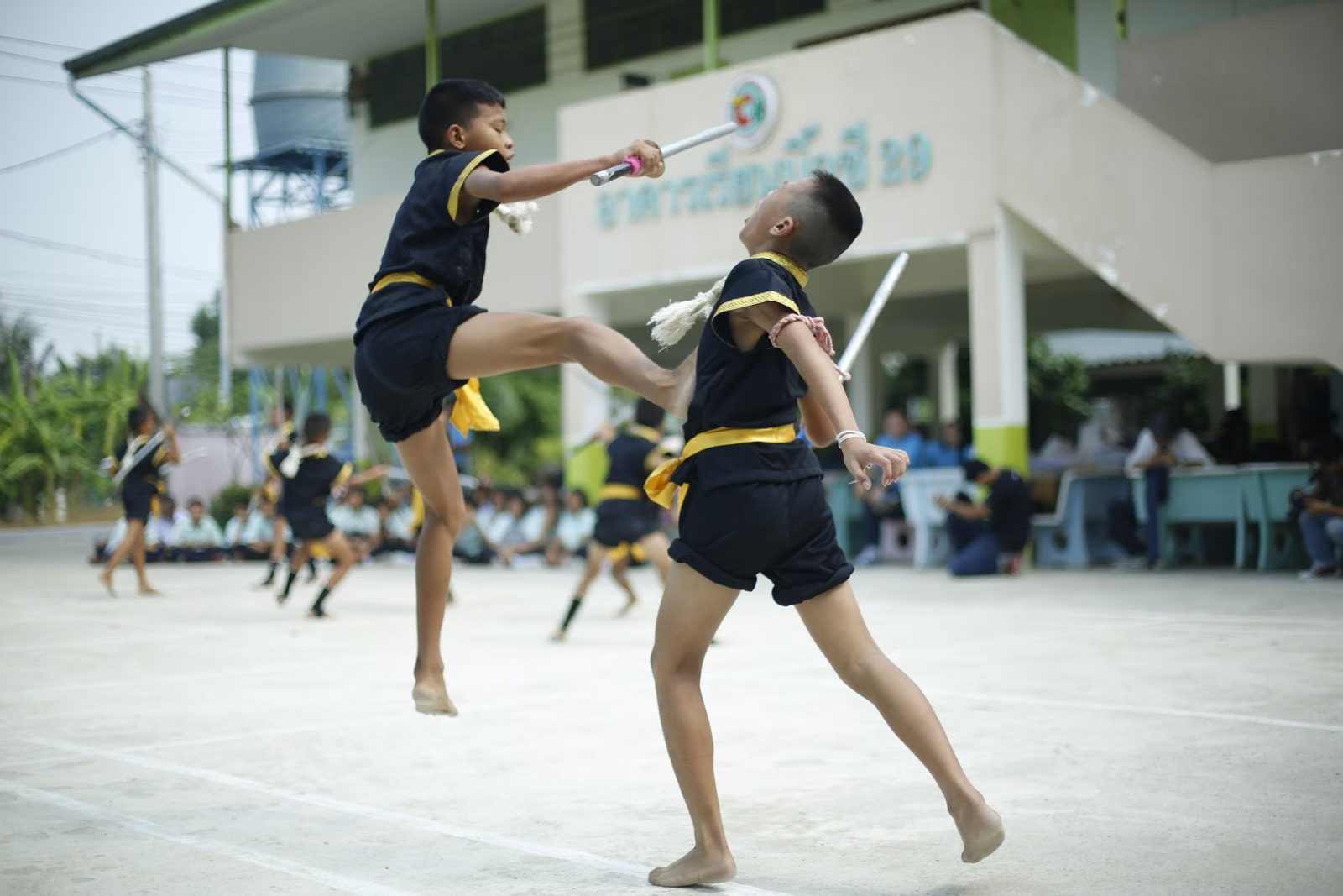 Thai schoolchildren in a martial arts display in a schoolyard, one mid-kick, with classmates watching from the shade