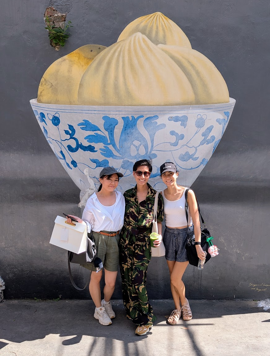 Three friends posing in front of a large blue-and-white bowl mural on Song Wat, Bangkok
