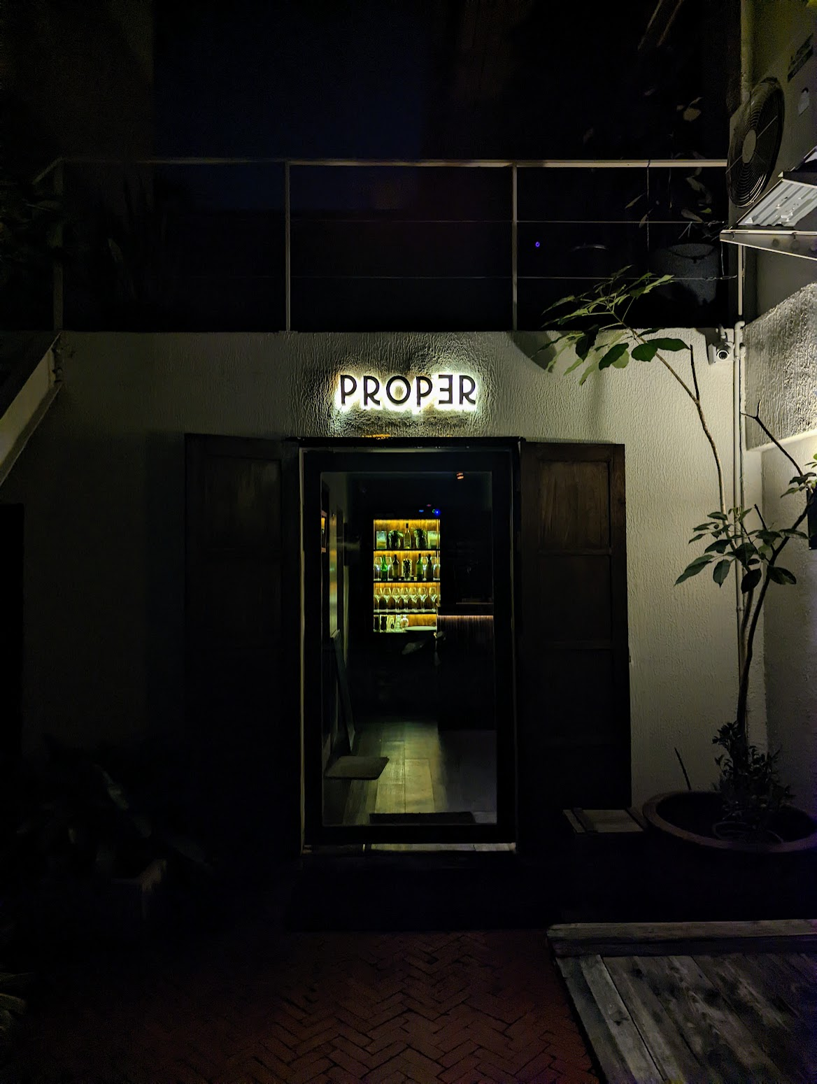 The dimly lit entrance to Proper bar at night, its name glowing above a doorway that reveals a backlit shelf of bottles and glassware